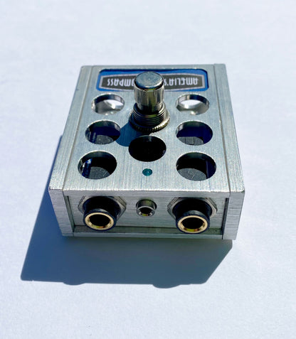 A metal foot switch with multiple input jacks and a central button, with the brand 'Amelia's Compass' visible on the top surface.