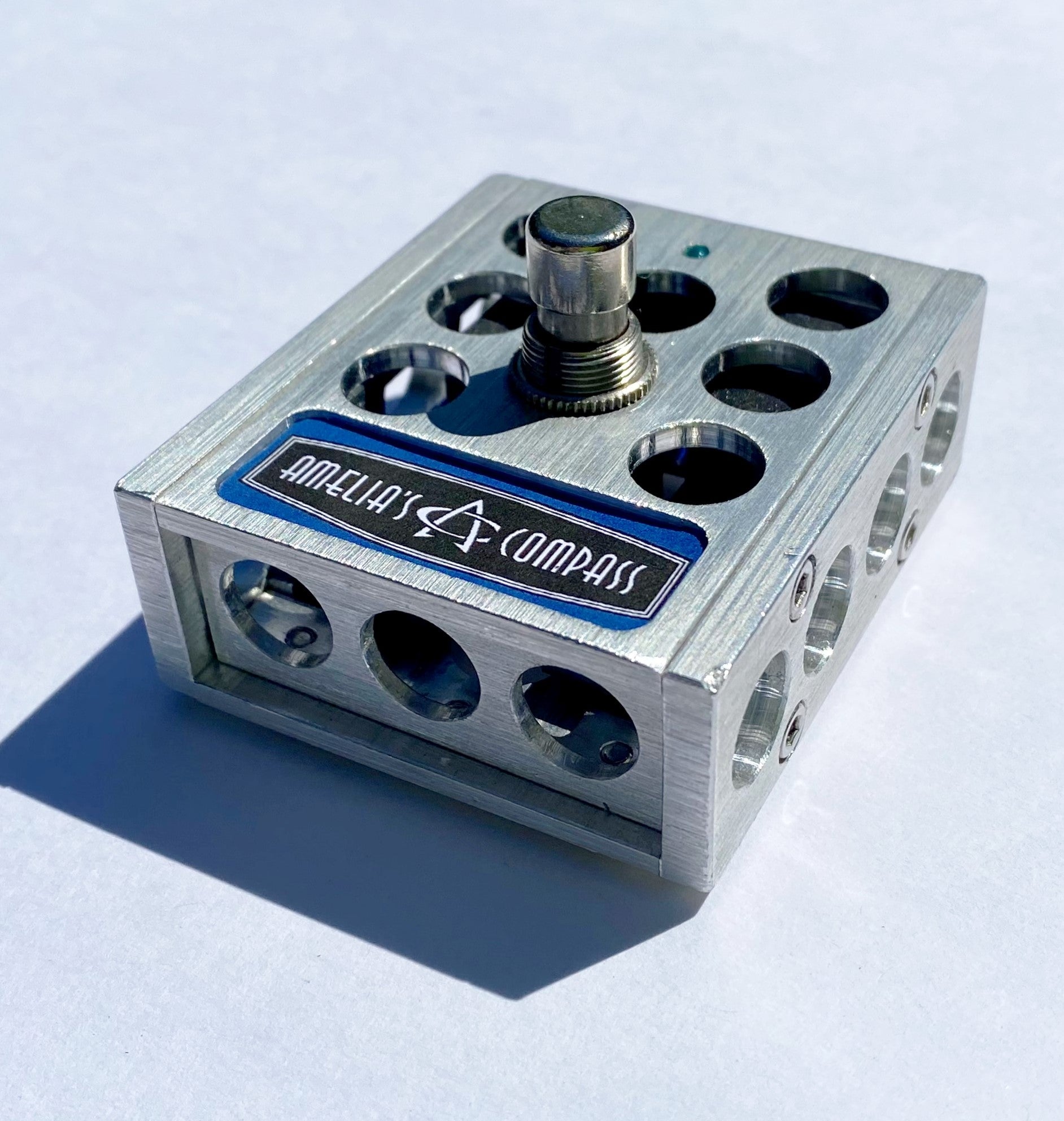 A metal foot switch with multiple input jacks and a central button, with the brand 'Amelia's Compass' visible on the top surface.